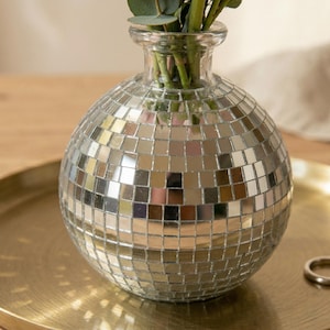 May include: A round, silver disco ball vase with a clear glass neck, filled with green foliage. The vase is made of small, mirrored squares. The vase sits on a gold-colored tray.