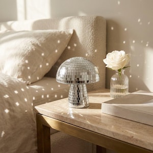 May include: A silver mushroom-shaped lamp with a mirrored surface sits on a marble-topped table. A white rose in a small glass vase and a closed book are also on the table. The lamp is next to a bed with a beige headboard and pillows.