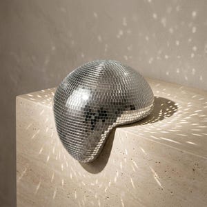 May include: A silver disco ball with a unique, asymmetrical shape. The surface is covered in small, reflective squares that catch the light, creating a dazzling effect. The disco ball is placed on a light-colored, textured surface.