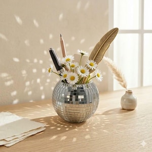 Könnte beinhalten: Eine silberne Kugelvase mit Mosaikmuster enthält Gänseblümchen, eine Feder, einen Bleistift und einen Stift. Sie steht auf einer Holzoberfläche, rechts daneben eine kleine cremefarbene Vase. Links liegt ein Stapel Papier.
