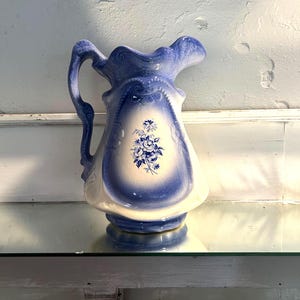 May include: A vintage blue and white ceramic pitcher with a floral design. The pitcher has a curved handle and a flared spout. The body of the pitcher is decorated with a blue floral pattern and a white background. The pitcher is sitting on a glass shelf.