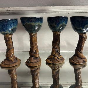 Puede incluir: Cuatro copas de cerámica hechas a mano. Cada copa tiene un cuenco esmaltado azul oscuro y un tallo marrón texturizado que se asemeja a ramas de árboles. Las copas se exhiben sobre una superficie reflectante, mostrando su diseño único.