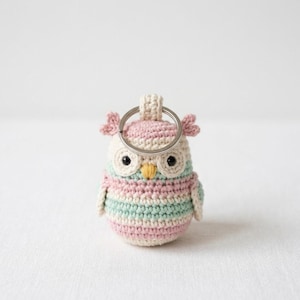 以下が含まれることがあります： パステルカラーの手編みフクロウのキーホルダー。フクロウは、ピンクのアクセントが付いたクリーム色の頭、大きな黒い目、黄色のくちばしが特徴です。体はピンクとミントグリーンの水平ストライプです。シルバーのキーリングが付いています。