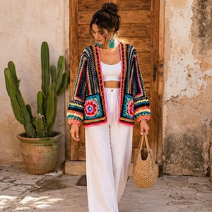 Op de afbeelding: Een veelkleurig gehaakt vest met een granny square patroon in levendige tinten roze, blauw, geel en zwart. Het vest heeft lange mouwen en wordt gedragen over een witte crop top en een witte wijde broek, gecombineerd met een gevlochten tas.
