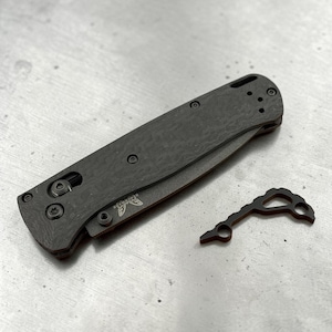 Puede incluir: Una navaja plegable cerrada con mango de fibra de carbono oscuro y texturizado con tornillería negra, que presenta el logotipo de Benchmade impreso en la hoja. Junto a la navaja hay una pequeña herramienta metálica negra angular con un recorte.