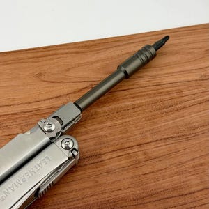 May include: A silver multi-tool with a dark grey screwdriver attachment. The multi-tool is partially open, showing the Leatherman brand name. The background is a wooden surface.