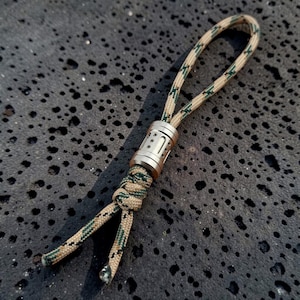 May include: A tan and green patterned paracord lanyard featuring a cylindrical metallic bead with vertical grooves and small circular indentations. The cord is looped through the centre of the bead and secured with a knot at the bottom end.