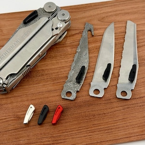 May include: A multi-tool with the text "LEATHERMAN WAVE+" and several interchangeable blades and tools. The blades include a saw, a knife, and a hook blade. There are also three small, coloured inserts: silver, black, and red. All items are on a wooden surface.