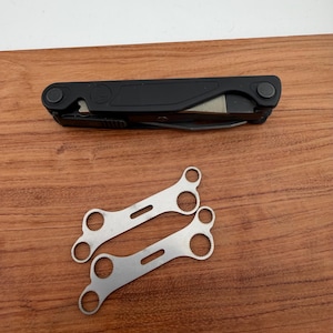 May include: A matte black multi-tool in a folded position featuring a circular logo on the handle, accompanied by two flat, silver-coloured metal wrenches with multiple serrated circular openings of varying sizes arranged along their length.