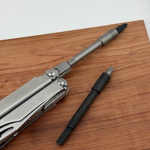 May include: A silver multi-tool, marked "LEATHERMAN WAVE+", with two attachments. One attachment is black, the other silver. The items are displayed on a wooden surface.