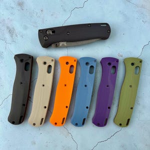 May include: A collection of six knife handle scales in black, beige, orange, blue, purple, and olive green, arranged in a row below a single folded black pocket knife with a visible metal blade and a Benchmade logo on the pivot area.