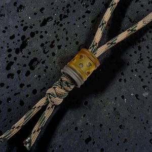 May include: A tan and green camouflage patterned paracord lanyard featuring a cylindrical bead with a translucent amber-coloured top section and a solid grey base. The cord is tied in a decorative knot below the bead, securing the two ends together.