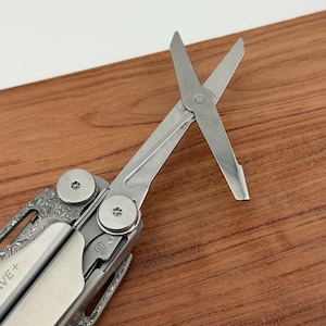 May include: A silver multi-tool with open scissors. The handle has a patterned design. The scissors blades are open. The background is a wooden surface.