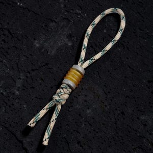 Peut inclure: Une lanière en paracorde à motifs beiges et verts dotée d'une perle cylindrique centrale avec un centre ambré translucide et des extrémités en métal argenté texturé, fixée par un nœud décoratif au bas de la boucle.