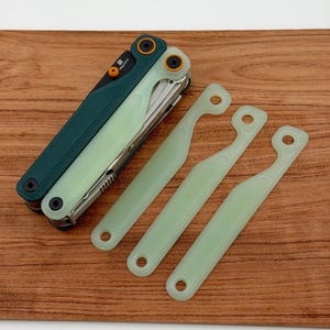 Könnte beinhalten: Ein Multitool mit dunkelgrünem Griff und hellgrüner Klinge, zusammen mit drei hellgrünen, gebogenen Werkzeugen. Das Multitool hat orangefarbene Akzente und den Text "Magicut". Die Artikel sind auf einer Holzoberfläche angeordnet.