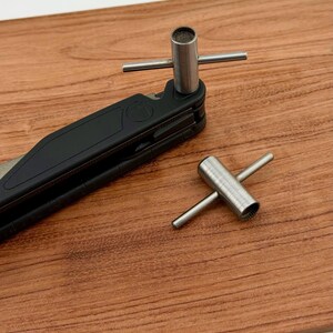 Könnte beinhalten: Ein schwarzes Multitool mit einem silbernen Metallbit und einem separaten silbernen Metallwerkzeug auf einer Holzoberfläche. Das Multitool ist teilweise geöffnet und zeigt seine Komponenten. Das silberne Werkzeug hat einen zylindrischen Kopf mit einem Quersteg.