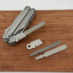 Könnte beinhalten: Ein silbernes Multitool mit der Aufschrift "LEATHERMAN WAVE" ist teilweise geöffnet und zeigt verschiedene Werkzeuge. Zwei Sägeblätter und ein Klingenhalter sind ebenfalls zu sehen, auf einer braunen Holzoberfläche.