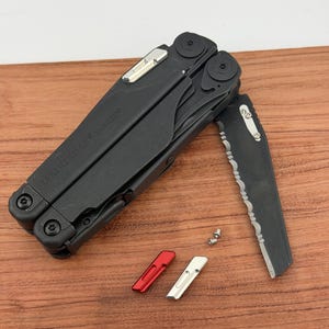 Pode incluir: Multi-ferramenta preta com o texto "LEATHERMAN SURGE" na lateral. A ferramenta tem uma lâmina parcialmente serrilhada, e duas pequenas inserções vermelhas e prateadas estão ao lado. A ferramenta está sobre uma superfície de madeira.