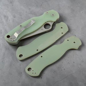 Op de afbeelding: Een inklapbaar zakmes met een lichtgroen handvat en een zilverkleurige metalen clip, vergezeld van twee extra bijpassende lichtgroene handgrepen. Het mes heeft een rond duimgat in het lemmet en zichtbare zilverkleurige schroeven.