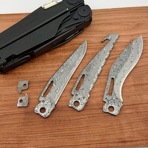Könnte beinhalten: Ein schwarzes Multitool mit mehreren austauschbaren Klingen und Teilen, darunter eine Sägeblatt und zwei kleine Teile. Die Klingen haben ein einzigartiges Muster. Die Artikel sind auf einer Holzoberfläche angeordnet.