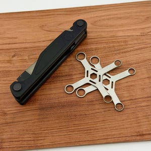 Op de afbeelding: Een zwarte multitool en twee zilveren multispannergereedschappen worden weergegeven op een houten oppervlak. De multitool is opgevouwen en heeft een strakke, matte afwerking. De multispaners hebben een stervormig ontwerp met meerdere openingen.