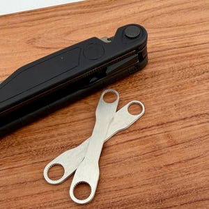 Peut inclure: Un outil multifonction noir avec deux clés en métal argenté. Les clés ont une ouverture circulaire à chaque extrémité. Les outils sont sur une surface en bois.