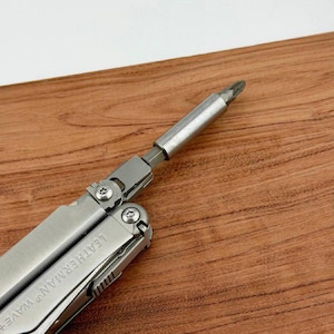 Puede incluir: Una multiherramienta plateada con una punta de destornillador adjunta. La herramienta está parcialmente abierta, revelando el logotipo de Leatherman Wave+. El fondo es una superficie de madera.