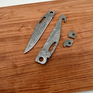 Pode incluir: Close-up de três lâminas de faca de metal e duas pequenas peças de metal com um design único. As lâminas têm uma ponta e uma ponta curva. As peças de metal têm um buraco no centro. Os itens estão em uma superfície de madeira.