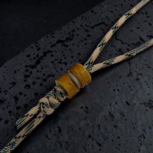 Op de afbeelding: Een lanyard gemaakt van beige, groen en zwart camouflage paracord met een doorschijnende, amberkleurige cilindrische kraal. Het koord is onder de kraal vastgeknoopt met een decoratieve knoop die de twee strengen van het koord bij elkaar houdt.