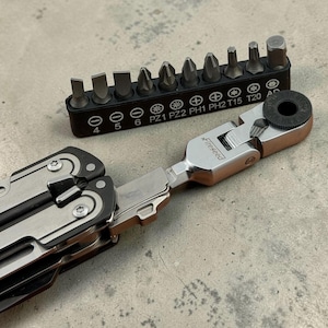Op de afbeelding: Een zilverkleurige en zwarte multitool met een inklapbare ratelkop met het opschrift Fireseed, naast een zwarte kunststof bithouder met tien diverse metalen schroefbits, waaronder platte, kruiskop- en Torx-maten.