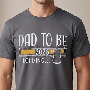 May include: Grey t-shirt featuring the text "DAD TO BE 2026 LOADING..." in white. The design includes a yellow loading bar and a baby bottle graphic. A casual tee for expectant parents.