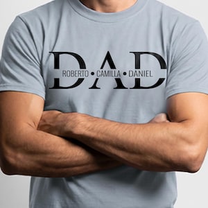 May include: A pale blue t-shirt featuring the word "DAD" in large, bold, black lettering. Beneath, the names "ROBERTO", "CAMILLA", and "DANIEL" are printed in smaller black text. The person in the image has their arms crossed.
