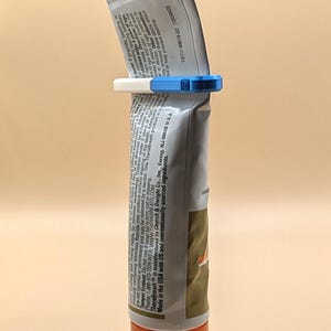 May include: A silver toothpaste tube with an orange cap, held upright by a blue and white plastic tube squeezer clip. The tube features printed text including TheraBreath, manufacturing information, and usage directions in English.