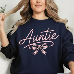 Op de afbeelding: Marineblauw sweatshirt met het woord "Auntie" in roze script, vergezeld van een roze strik. Het sweatshirt is effen van kleur en de tekst en strik zijn het middelpunt van het ontwerp.