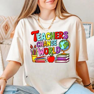 Könnte beinhalten: Cremefarbenes T-Shirt mit farbenfrohem Aufdruck "Teachers Change World". Das Design umfasst einen Globus, Bücher, Bleistifte, Pinsel und einen Apfel. Ein lässiges Kleidungsstück für den Alltag.