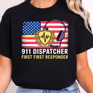 Puede incluir: Camiseta negra con un diseño que presenta la bandera estadounidense, un escudo con un gráfico de latido cardíaco y unos auriculares. El texto dice "911 DISPATCHER FIRST RESPONDER" en blanco y amarillo.
