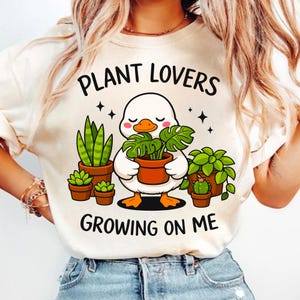 May include: Cream-colored sweatshirt featuring a cartoon duck holding a potted plant, surrounded by other potted plants. The text "PLANT LOVERS GROWING ON ME" is printed above and below the duck illustration. The sweatshirt is worn by a person.