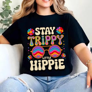 以下が含まれることがあります： 黒いTシャツ。カラフルなレトロフォントで「STAY TRIPPY LITTLE HIPPIE」と書かれています。虹色のレンズと花のアクセントが付いたサングラスのデザインで、1960年代または70年代を彷彿とさせます。