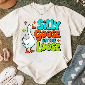 Peut inclure: T-shirt blanc avec un dessin d'oie de dessin animé et le texte "Silly Goose on the Loose". Le texte est en bleu, orange et vert, avec des étoiles. L'oie a un bec et des pattes oranges.