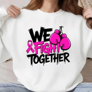 May include: White sweatshirt with the text "WE FIGHT TOGETHER" in black and pink. The design includes a pink ribbon and a pair of pink boxing gloves, symbolizing support and solidarity.