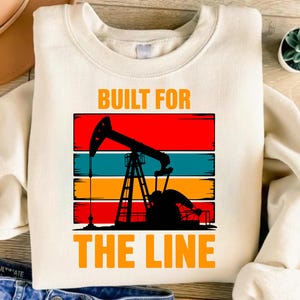 Puede incluir: Sudadera color crema con el texto "BUILT FOR THE LINE" en naranja. El diseño presenta una silueta de un balancín petrolero sobre un fondo de rayas rojas, turquesas, naranjas y negras.