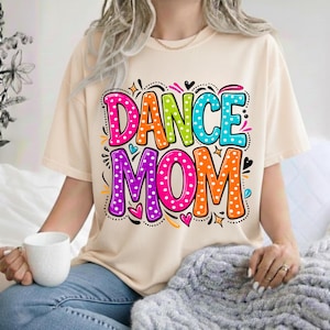 May include: A beige t-shirt with the words "DANCE MOM" in a colorful, polka-dotted design. The text is outlined in black and features hearts and stars. The shirt is worn by a person holding a white mug.