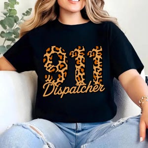 May include: Black t-shirt featuring the number "911" in a leopard print design, with the word "Dispatcher" in a cursive font below. The shirt is a crew neck style.