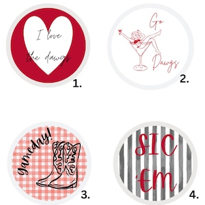 May include: A set of four circular buttons titled Georgia Buttons by XO By Kathryn Grace. Button 1 features a red heart with the text I love the dawgs. Button 2 shows a pin-up illustration in a martini glass with the text Go Dawgs. Button 3 displays cowboy boots on a red gingham background with the text Gameday!. Button 4 has black and white stripes with the red text SIC 'em.