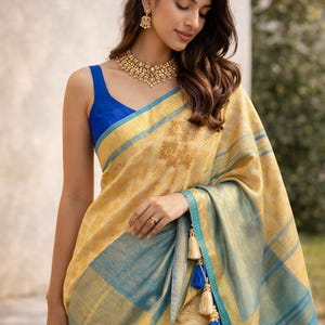 Könnte beinhalten: Eine Frau trägt einen gelb-blauen Sari mit Goldschmuck. Der Sari hat einen blauen Rand mit Quasten und den Text "KA'N'CHI ARTISAN SILKS by Laya" in der oberen linken Ecke. Der Sari hat ein goldenes Blumenmuster.