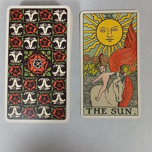 May include: A tarot card deck featuring the Sun card and the deck's back design. The Sun card depicts a person riding a white horse under a radiant sun. The deck's back has a repeating pattern of red and white floral designs on a dark background.