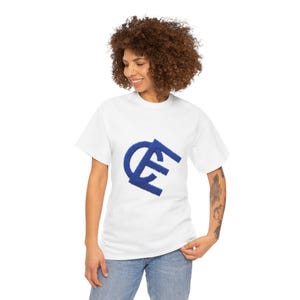 May include: A white t-shirt featuring a blue embroidered design. The design appears to be stylized initials or a logo. The shirt is made of a soft material and is being worn by a person.