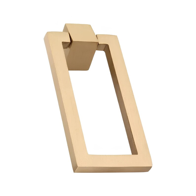 Puede incluir: Un tirador rectangular de color dorado con un dise&ntilde;o geom&eacute;trico. El tirador presenta un marco rectangular y un bloque de conexi&oacute;n. El herraje es de metal y est&aacute; sobre un fondo blanco.
