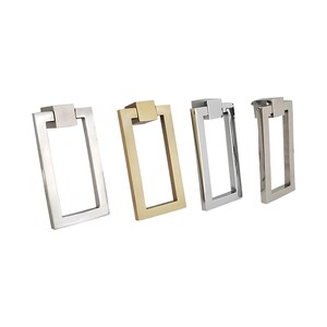 Puede incluir: Cuatro tiradores rectangulares para armarios en tonos plateados y dorados. Cada tirador presenta un asa rectangular con una parte superior cuadrada. Los tiradores est&aacute;n dispuestos en fila sobre un fondo blanco, mostrando su dise&ntilde;o y acabado.