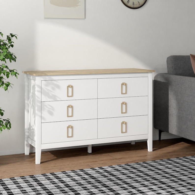 Puede incluir: Una c&oacute;moda blanca de seis cajones con tapa de madera clara y tiradores dorados. La c&oacute;moda est&aacute; colocada contra una pared blanca, con una planta y un sof&aacute; gris en el fondo. De estilo moderno.